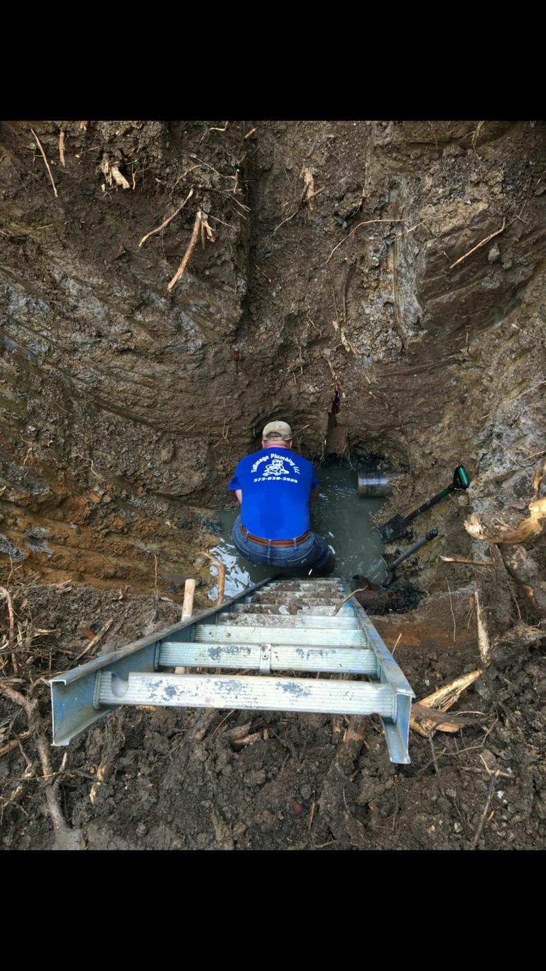 Looking down into a deep, wet excavation where a plumber in a Tallmage shirt is working at the bottom, with a ladder laid down into the hole