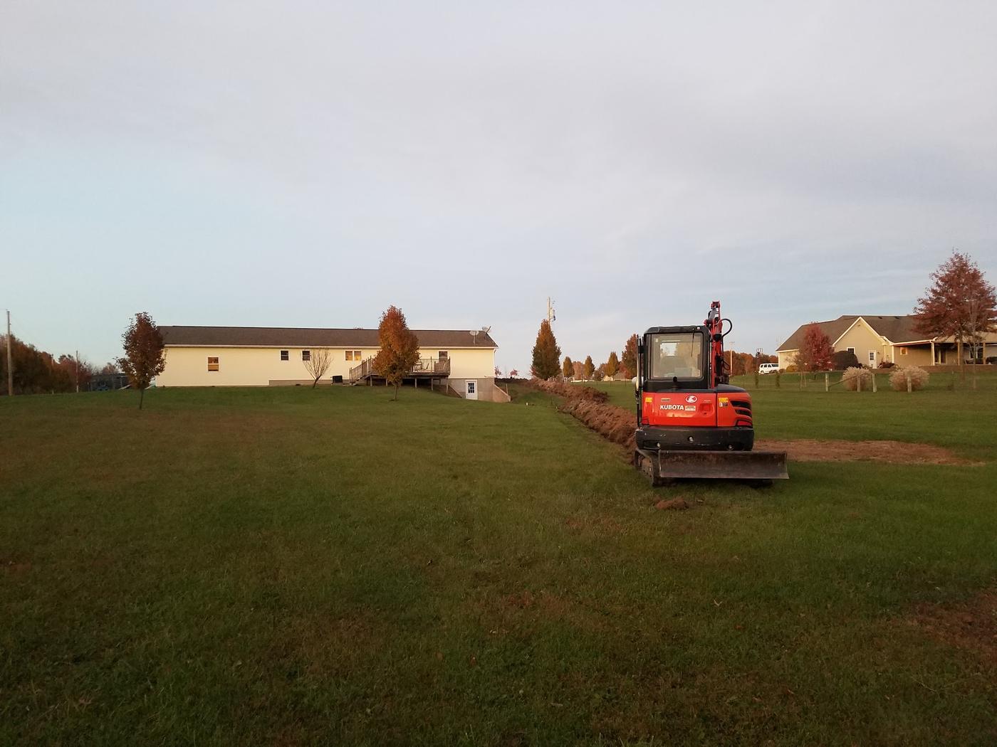 A tracked excavator at the end of a long trench cut across a lawn toward a house, at golden hour